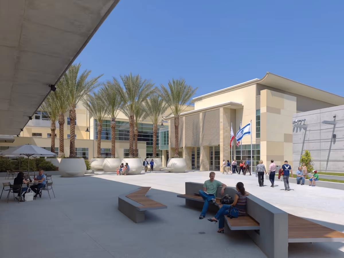 Outdoor courtyard area of a senior living facility with modern benches, large potted palm trees, and people sitting and walking around under a clear blue sky.