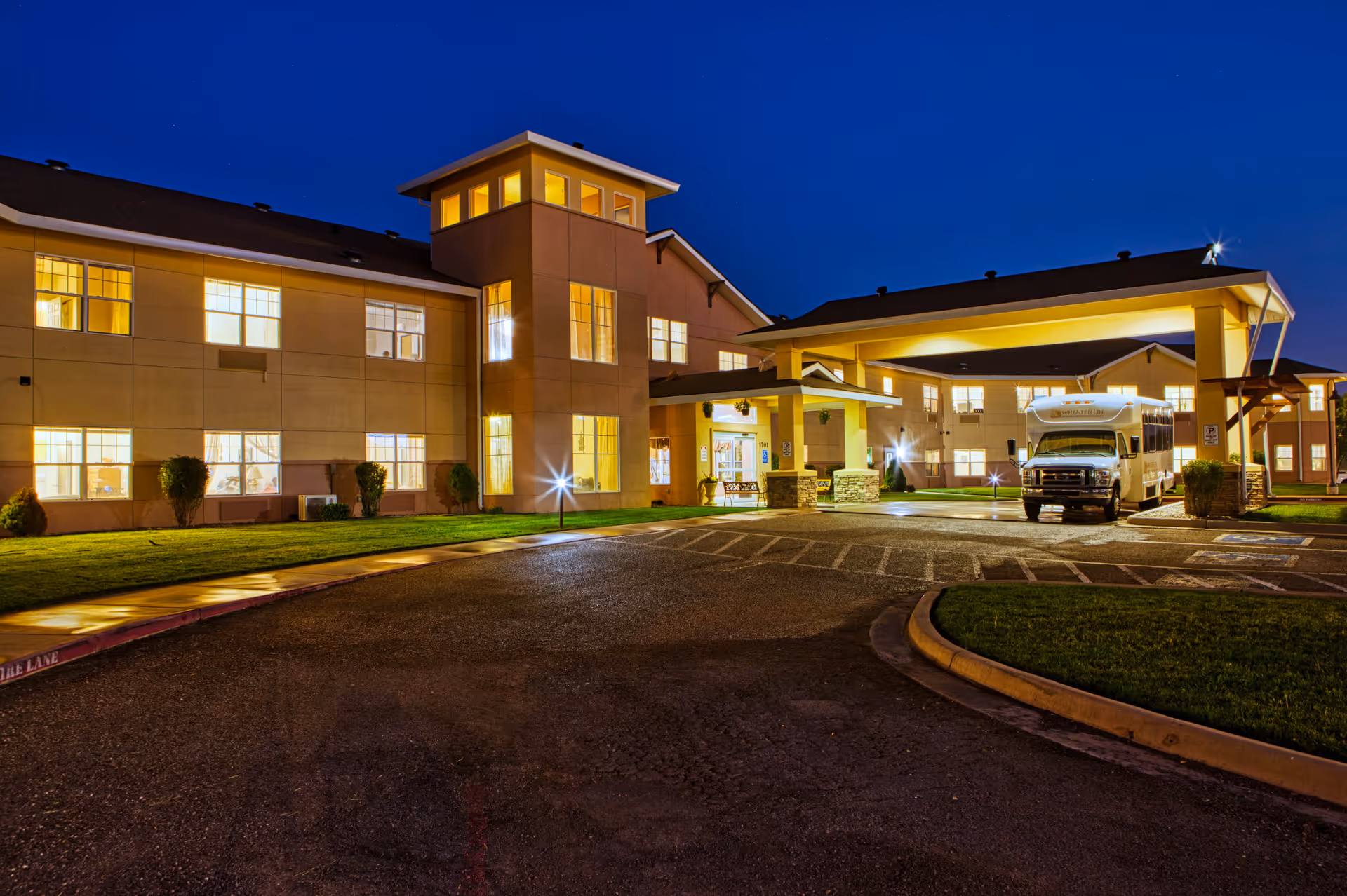 Exterior nighttime view of Wheatfields Estates Senior Living and Memory Care building with illuminated windows, a covered entrance, and a shuttle bus parked under the canopy.