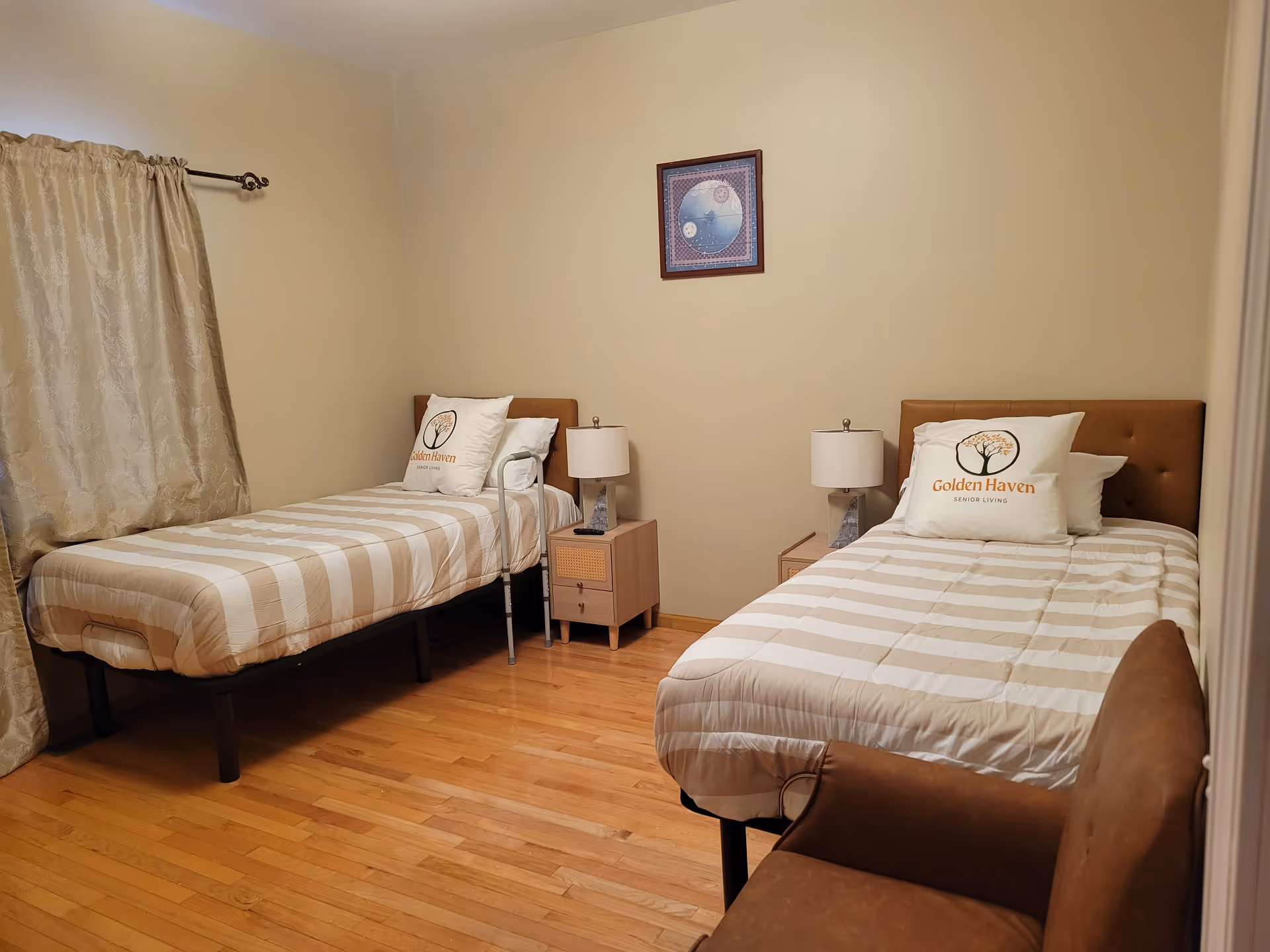 A bedroom with two single beds, each with beige and white striped bedding and a pillow with the Golden Haven Senior Living logo. There are two bedside tables with lamps between the beds. A brown armchair is partially visible in the foreground, and beige curtains cover a window on the left side. A framed picture hangs on the wall above the beds.