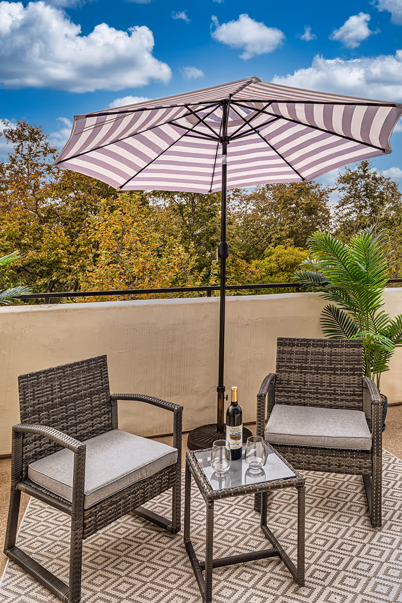 Outdoor patio area with two wicker chairs with gray cushions, a small glass-top table holding a bottle of wine and two glasses, a large striped umbrella providing shade, and green plants in the background under a blue sky with scattered clouds.