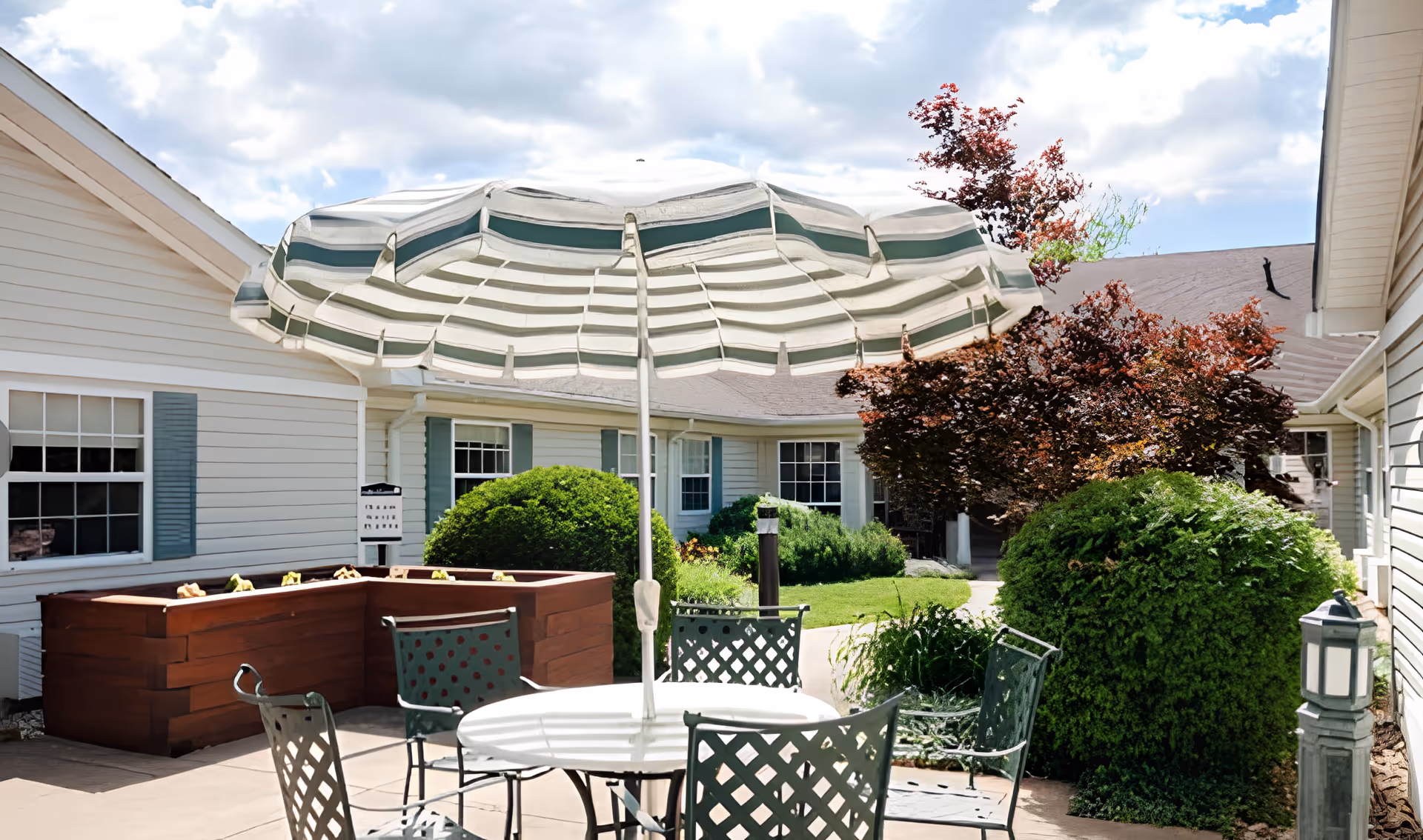 Outdoor patio area with a round white table and four metal chairs with lattice backs. A large striped umbrella shades the table. Surrounding the patio are green bushes, a raised wooden planter box, and white buildings with blue shutters. The sky is partly cloudy.