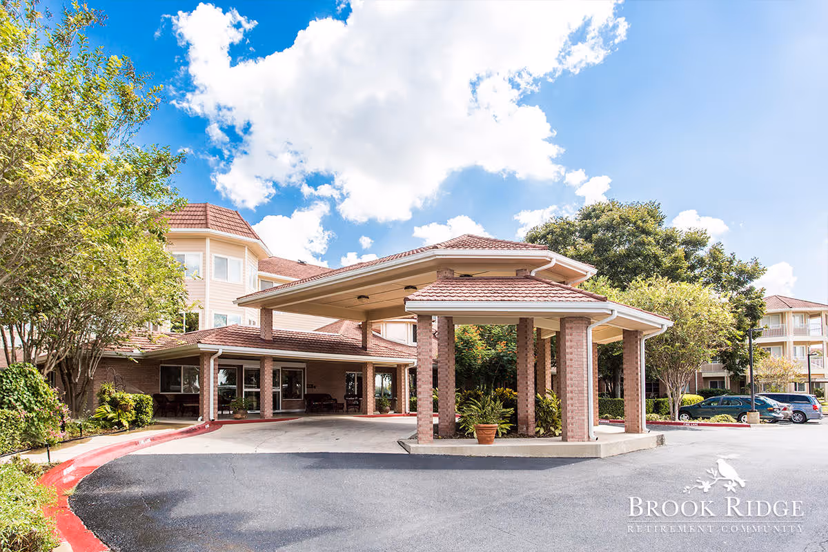 Covered porte-cochère and front entrance of Brook Ridge Retirement Community with landscaping, driveway, and parked cars under a blue sky.