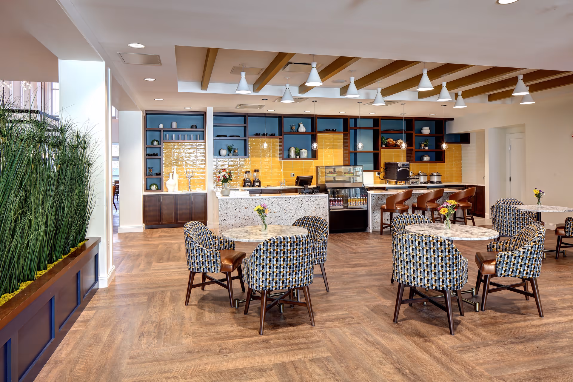 A bright and modern dining area with several round tables surrounded by patterned upholstered chairs. The back wall features a counter with bar stools, a display case with beverages, and shelves with decorative items. The floor has a wood pattern, and there are pendant lights hanging from the ceiling. A planter with tall green plants is visible on the left side.