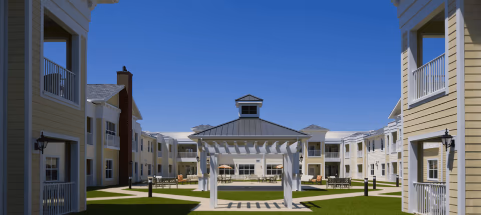 Sunny courtyard with a central white pergola, patio seating, and surrounding two-story yellow buildings with balconies under a clear blue sky.