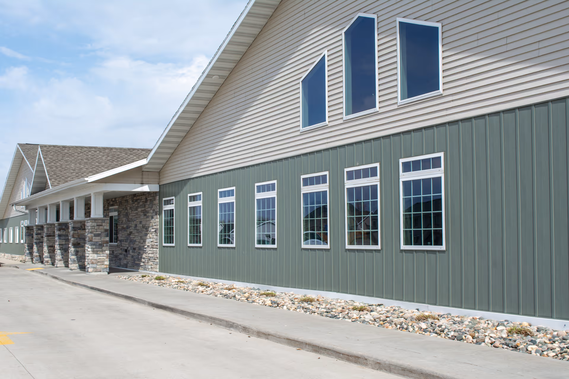 Exterior view of a building with green vertical siding and stone pillars supporting a covered entrance. The building has multiple rectangular windows and three triangular windows near the roofline under a partly cloudy sky.
