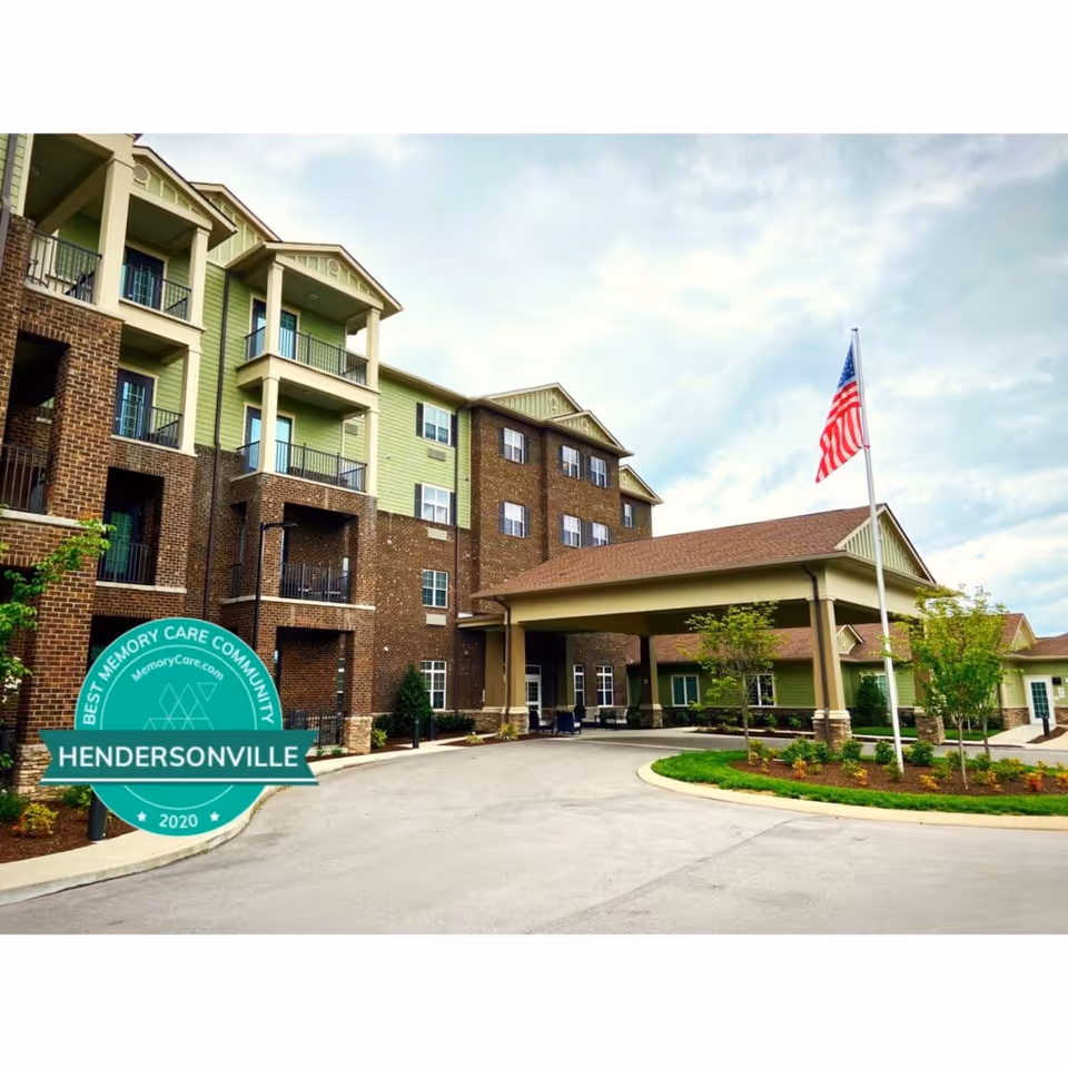 Exterior view of a multi-story senior living facility with green and brown brick facade, balconies, a covered entrance, landscaped greenery, and an American flag on a flagpole.