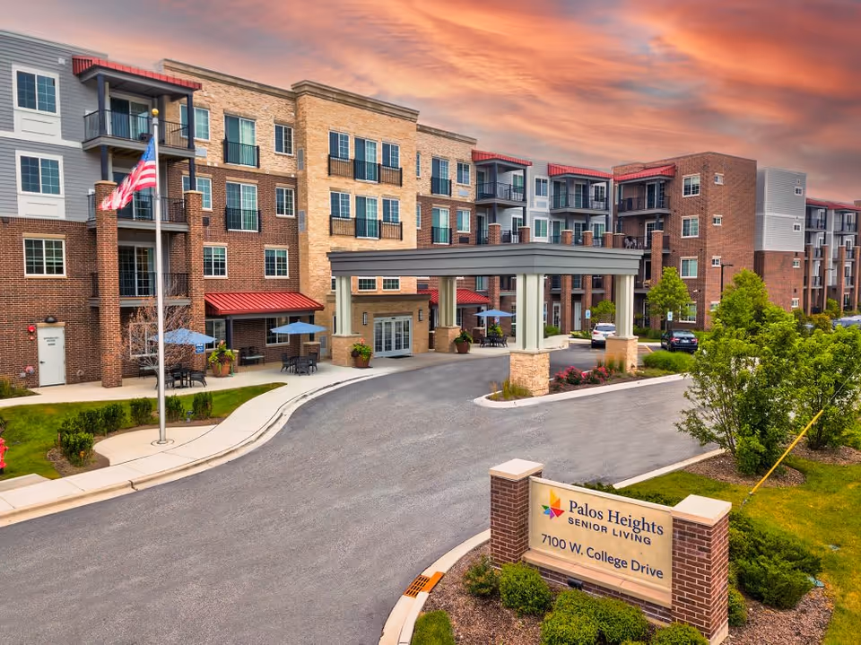 Exterior view of Palos Heights Senior Living facility at sunset, showing a multi-story building with balconies, an American flag on a flagpole, outdoor seating with umbrellas, a covered entrance driveway, and a sign with the facility name and address 7100 W. College Drive.