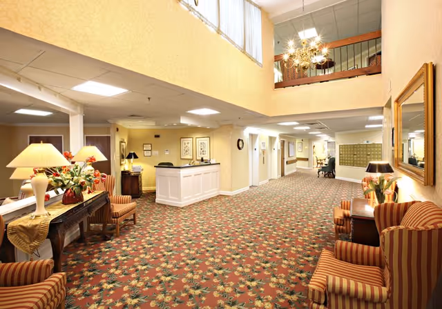 A spacious and well-lit interior hallway of a senior living facility with floral patterned carpet, a white reception desk, striped armchairs, lamps, and decorative plants. The hallway has multiple doors, a railing on the upper level, and a large mirror on the wall.