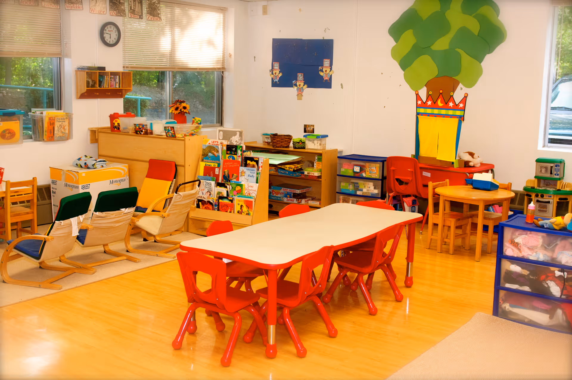 A brightly lit room with child-sized furniture including a white table with red chairs, small wooden chairs, and shelves filled with books, toys, and educational materials. The walls have colorful decorations including a large tree with a crown. There are windows letting in natural light and a clock on the wall.