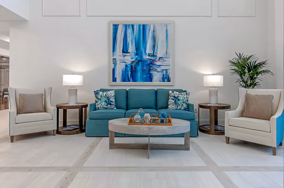 A modern living room with a teal sofa adorned with two floral patterned pillows, flanked by two beige armchairs each with a beige cushion. Two round wooden side tables with white lamps are positioned beside the sofa. A large abstract blue and white painting hangs on the white wall above the sofa. A round wooden coffee table with decorative items is centered in front of the sofa, and a green potted plant is placed behind the right armchair.