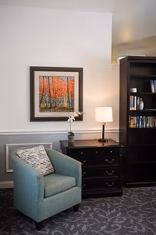 Cozy interior seating area with a teal armchair and patterned pillow beside a dark side table and lamp, a framed autumn painting on the wall, and a tall bookshelf.