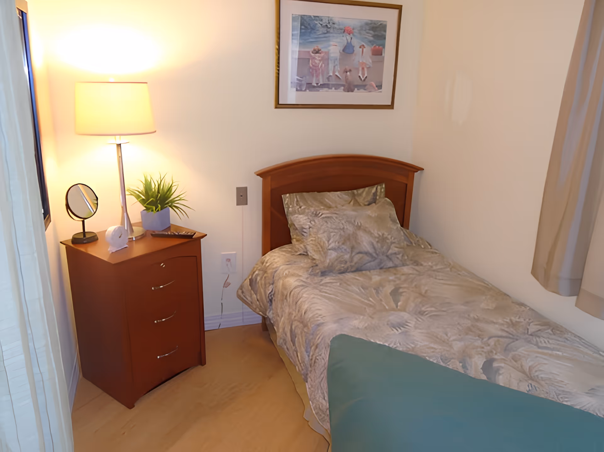A small bedroom with a single bed covered in a patterned bedspread and matching pillows. Next to the bed is a wooden nightstand with three drawers, holding a table lamp, a small round mirror, a decorative plant, and a remote control. A framed painting hangs on the wall above the bed, and beige curtains partially cover a window on the right side.