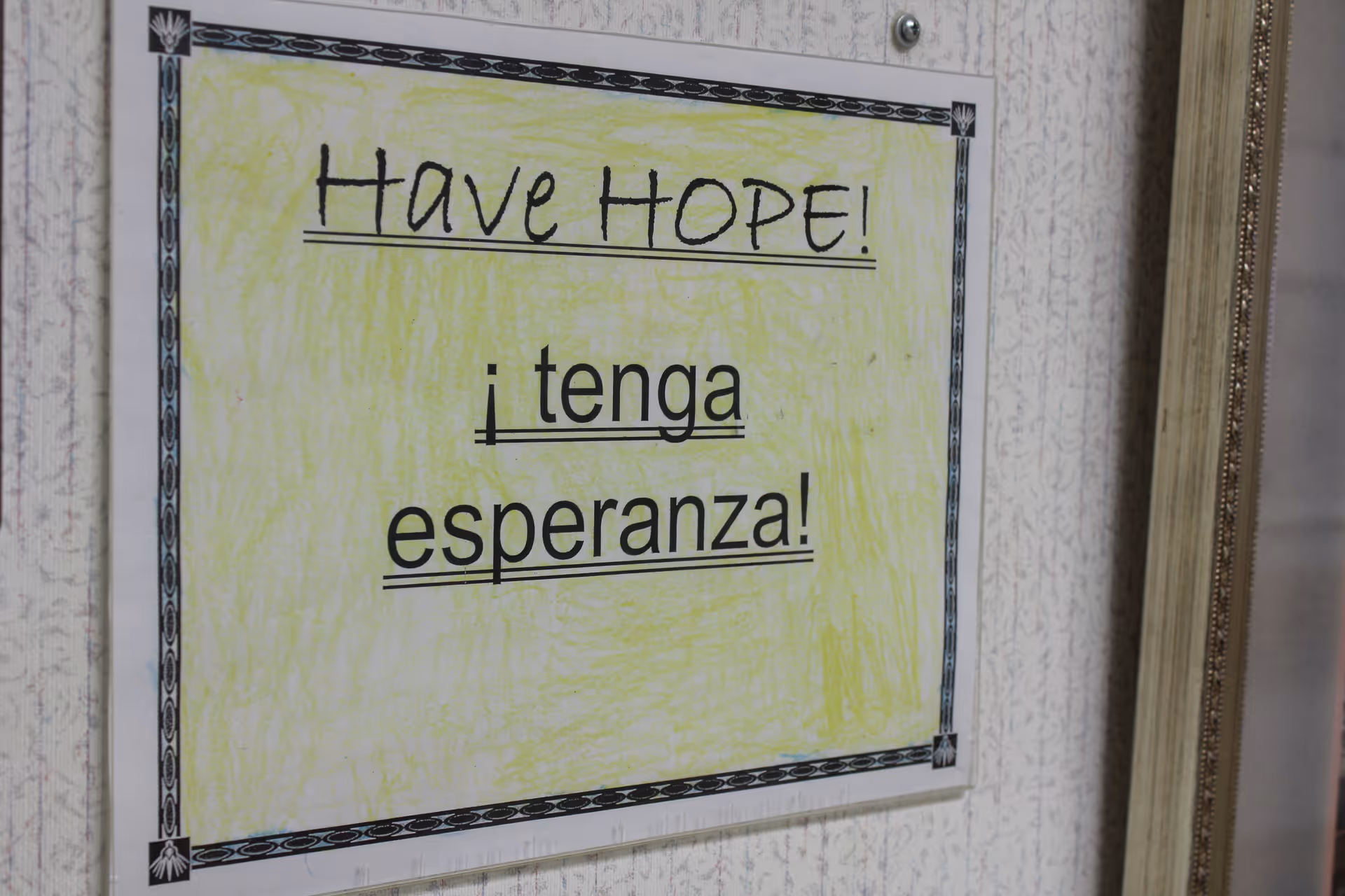 A framed sign on a wall with a yellow background and a decorative black border that reads 'Have HOPE!' and below it in Spanish '¡tenga esperanza!'