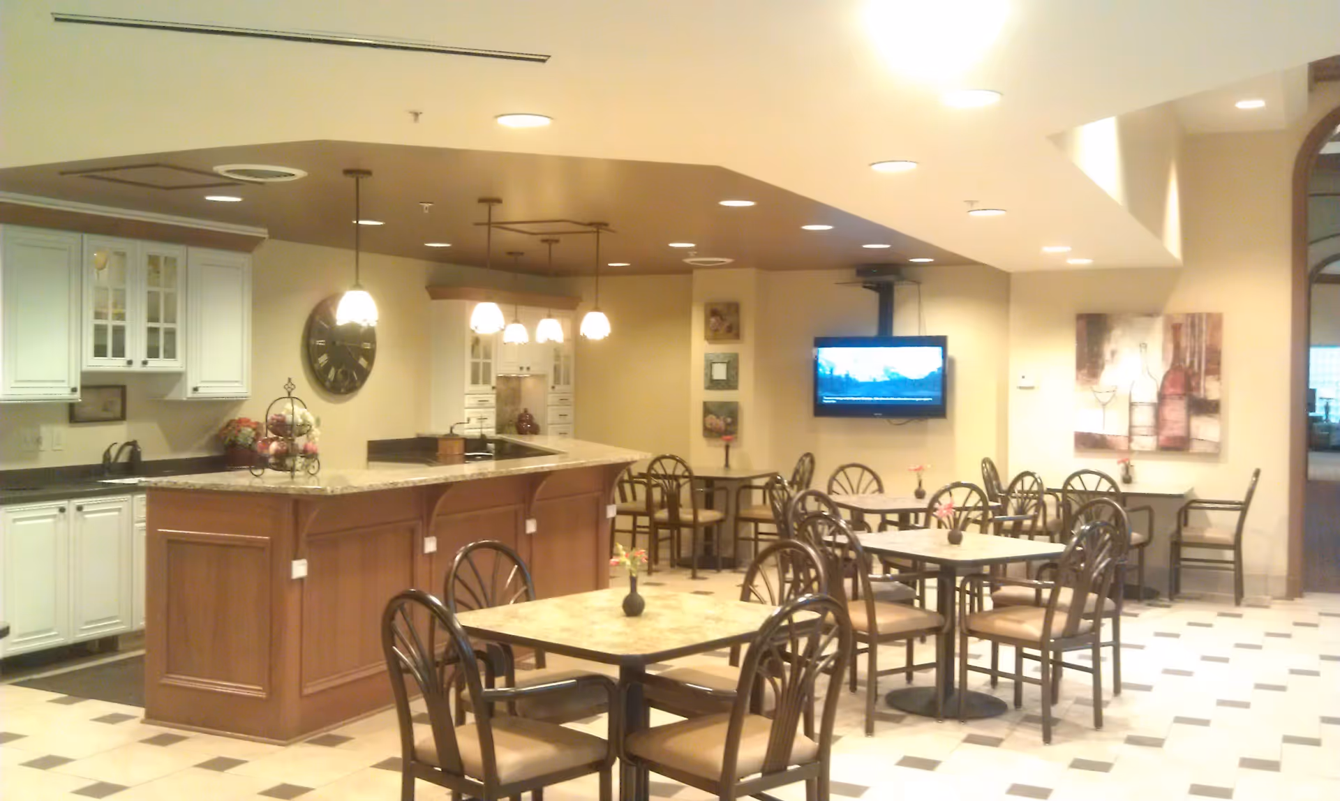 A dining area in a senior living facility featuring several tables with chairs, a kitchen counter with hanging pendant lights, white cabinets, a wall clock, and a wall-mounted television. The room has a tiled floor and warm lighting with decorative artwork on the walls.