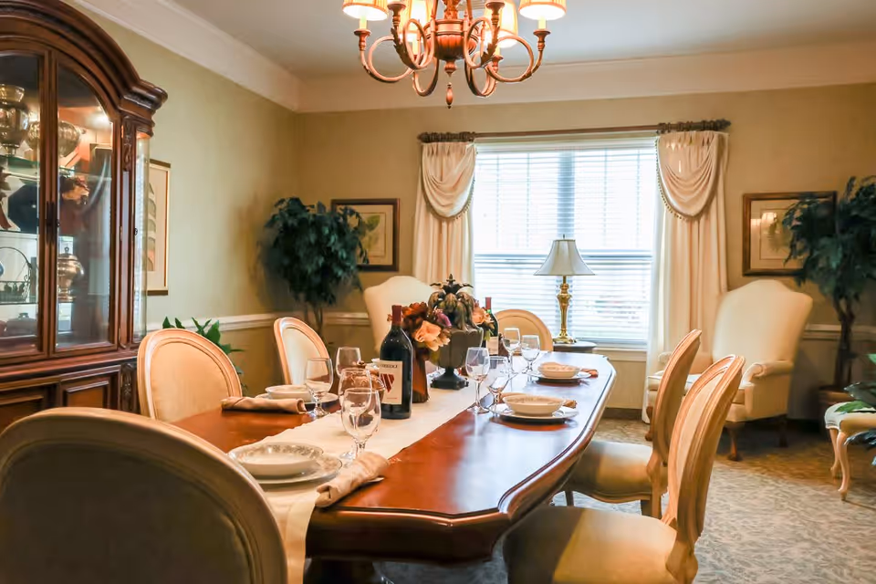 A formal dining room with a long wooden table set for six people. The table is adorned with a white runner, plates, bowls, wine glasses, napkins, and a centerpiece with flowers and a bottle of wine. The room has beige walls, a large window with draped curtains, two armchairs, a wooden china cabinet displaying glassware, and a chandelier hanging from the ceiling.