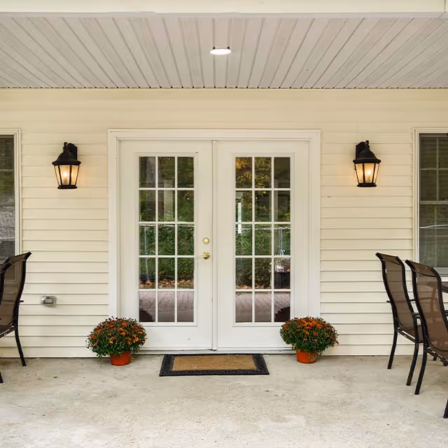 Exterior view of a building entrance with white double French doors flanked by two wall-mounted lantern-style lights. Two potted plants with orange flowers are placed symmetrically on either side of a doormat in front of the doors. There are two black chairs visible on each side of the entrance.