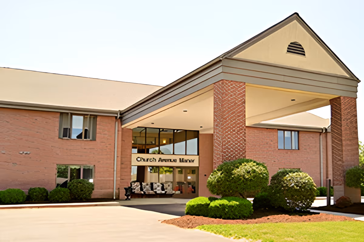 Front entrance of a brick senior living facility with a covered porte-cochere, glass doors, and trimmed bushes.