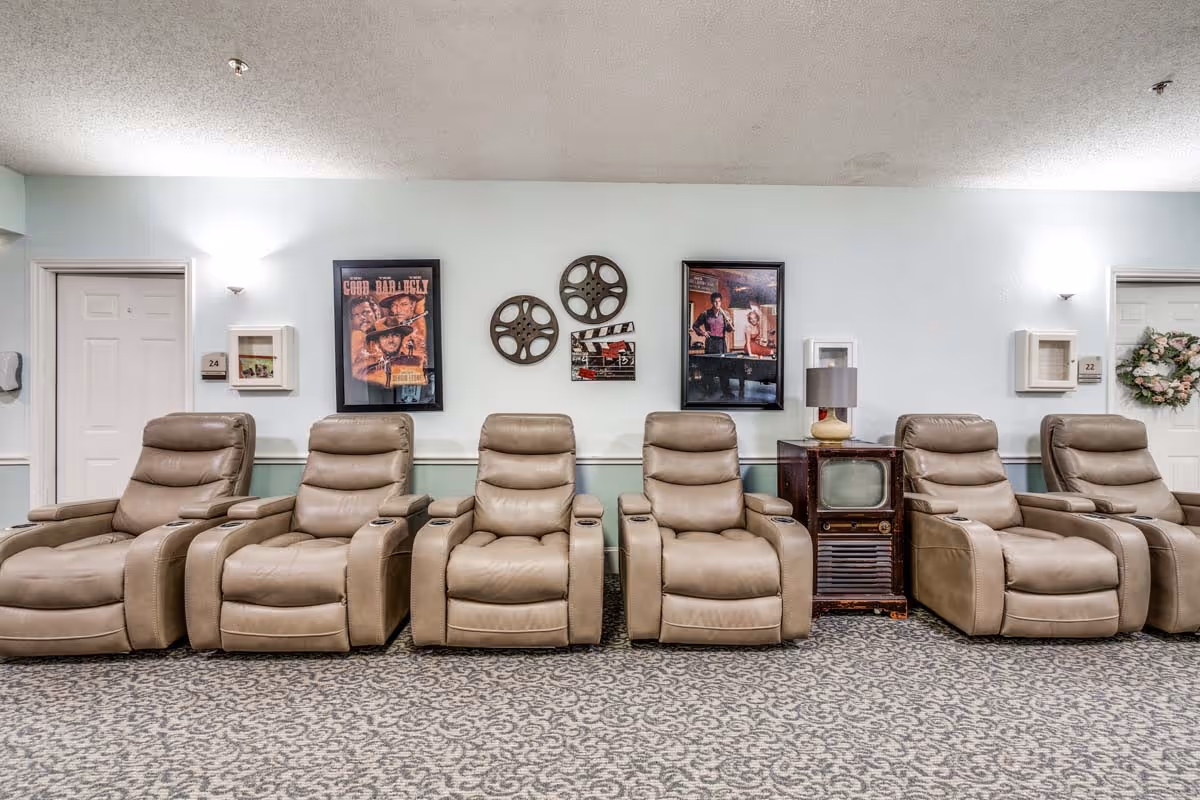 A row of six beige leather recliner chairs arranged in a common area with a patterned carpet. On the wall behind the chairs are two framed movie posters, two decorative film reels, and a clapperboard. There is an old-fashioned television set with a lamp on top between the fourth and fifth chairs. Two white doors with room numbers 24 and 22 flank the seating area, one door decorated with a floral wreath.