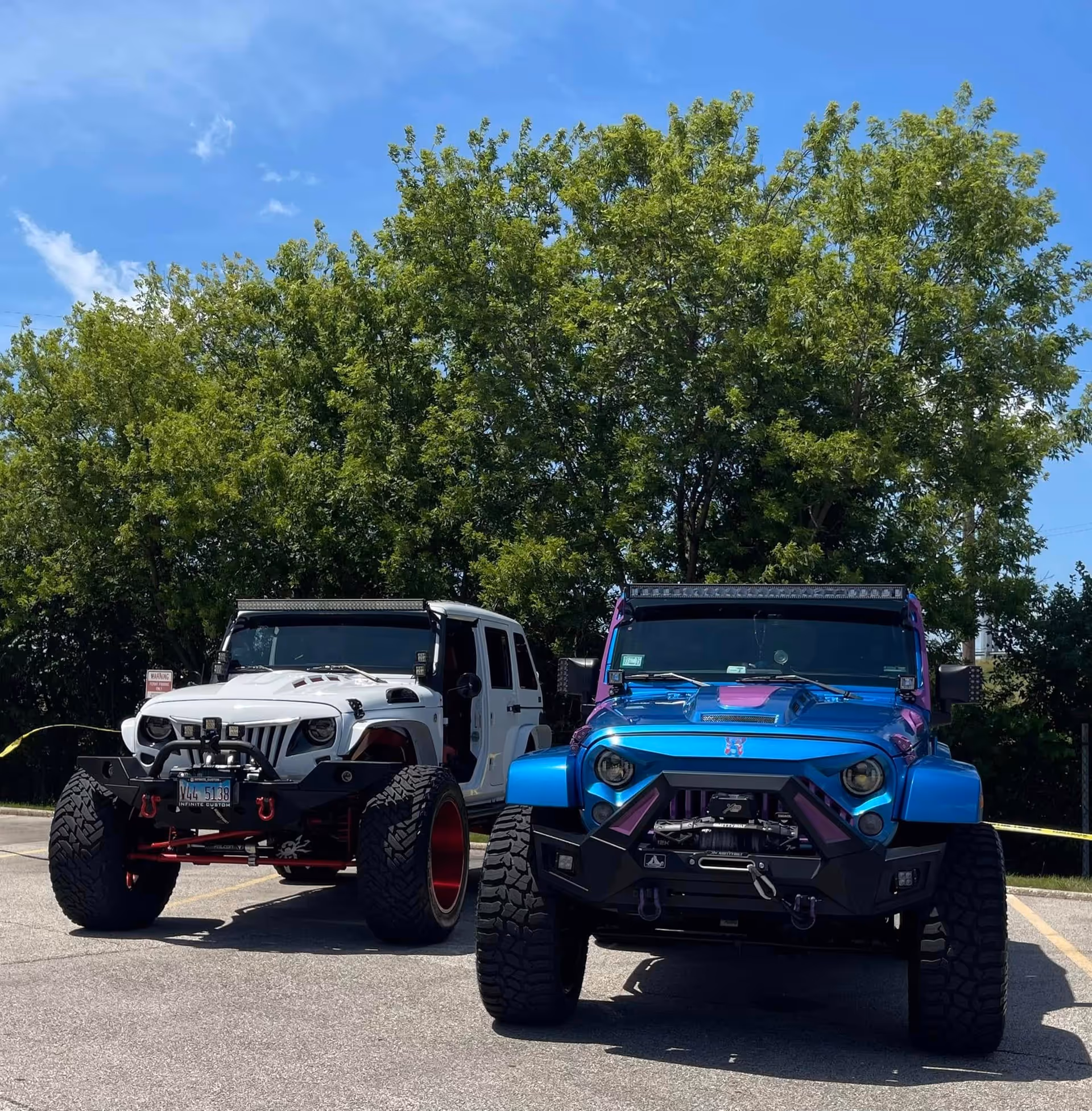 Two heavily modified off-road jeeps, one white and one blue, parked in a lot in front of large green trees under a blue sky.