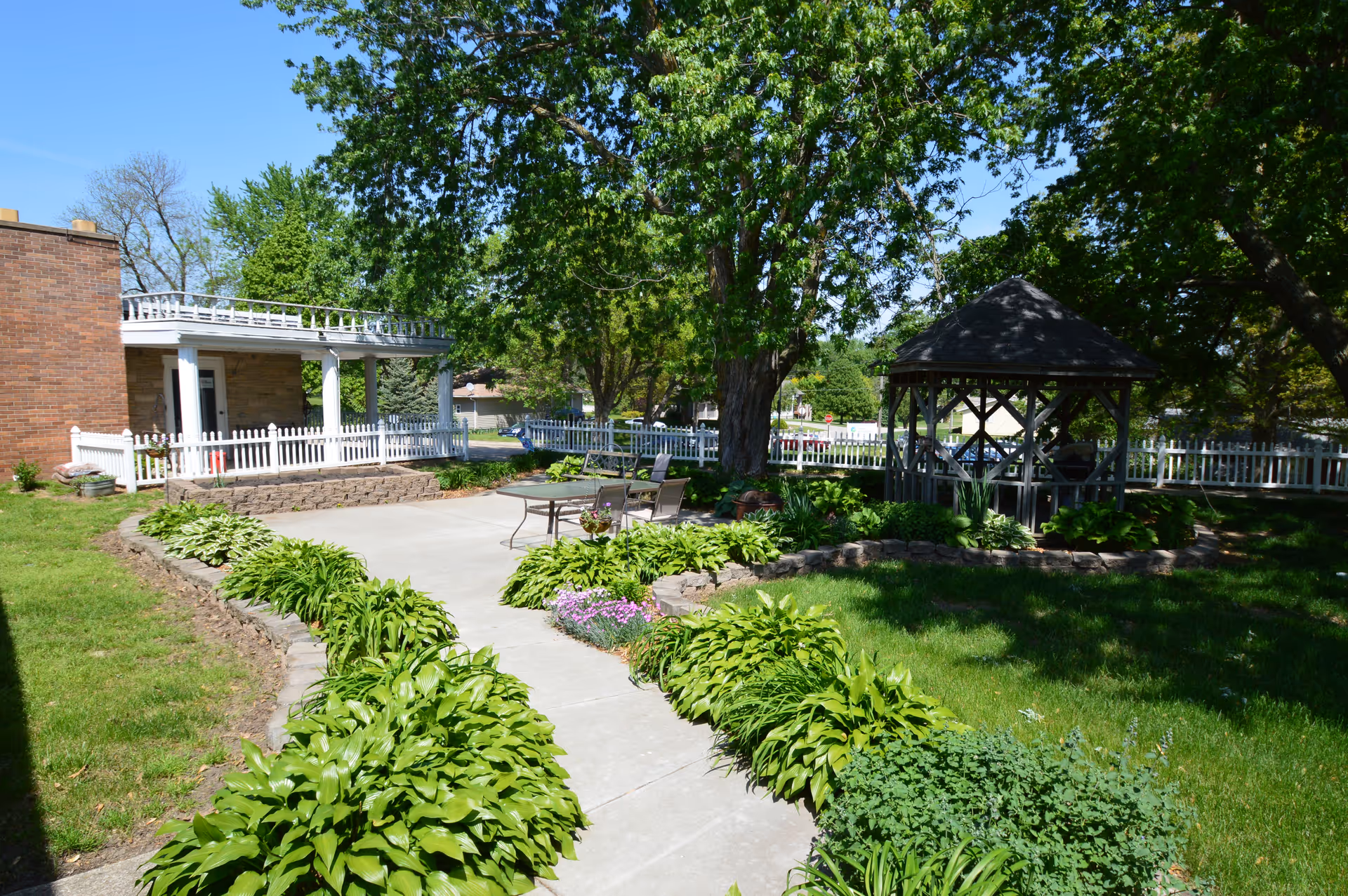 Landscaped outdoor courtyard with a concrete walkway, leafy plants, a gazebo, and patio seating beside a white picket fence and brick building.