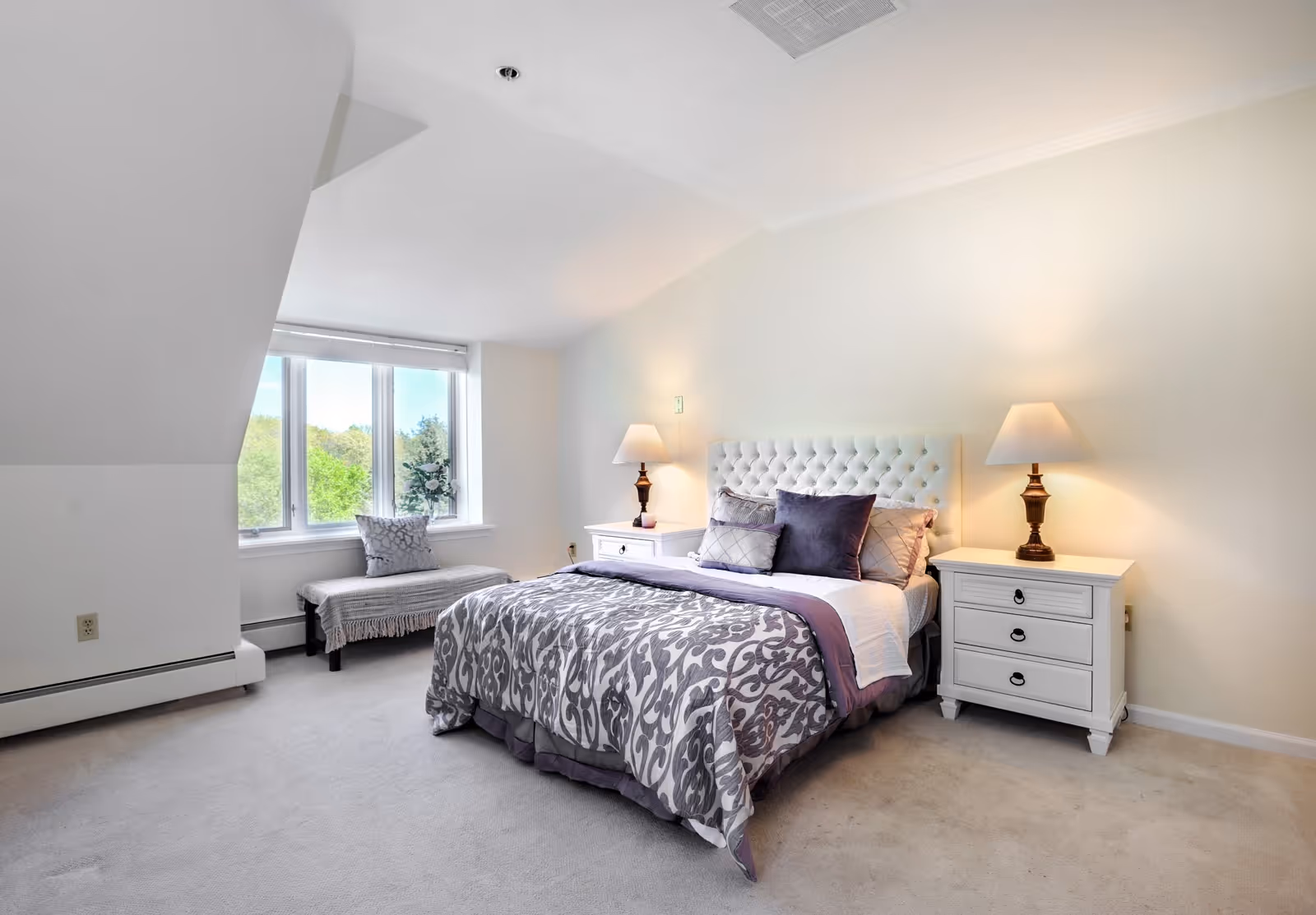 Bright bedroom with a tufted headboard bed, patterned bedding, two nightstands with lamps, and a window bench beneath large windows.