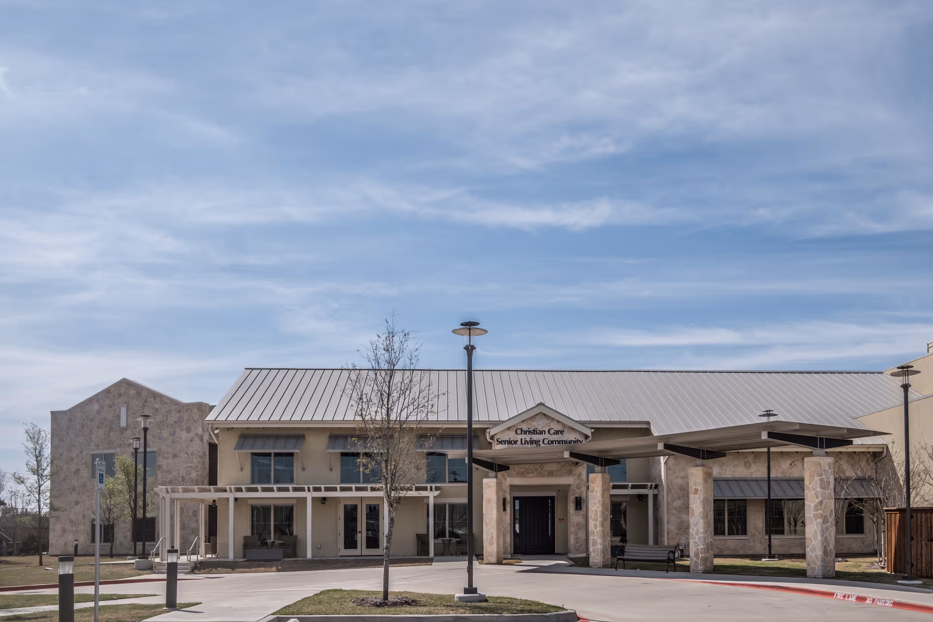 Exterior view of a senior living community building named Christian Care Senior Living Community with a stone facade, large windows, and a covered entrance. The sky is clear with some clouds, and there are a few young trees and lamp posts in front of the building.