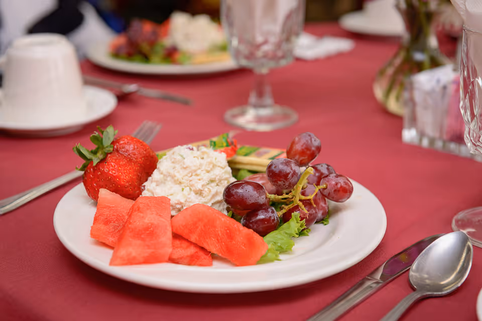 A white plate with watermelon, strawberries, grapes and a scoop of salad on a red-tablecloth dining table with glassware and cutlery.
