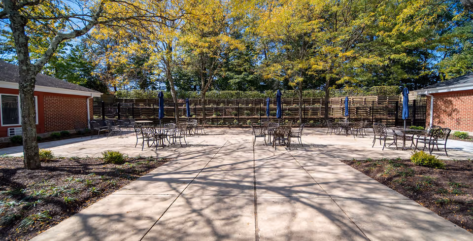 Outdoor patio area at Independence Court of Hyattsville with several metal tables and chairs, some with closed blue umbrellas. The patio is surrounded by trees with green and yellow leaves, and two red brick buildings are visible on either side.