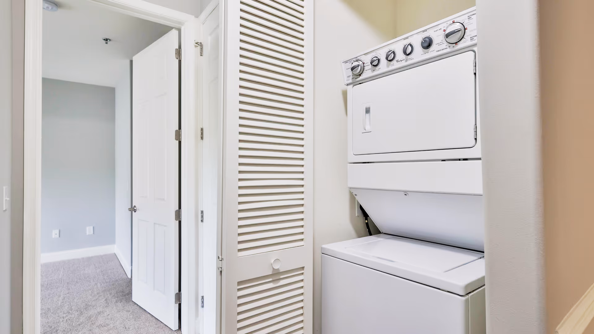 A small laundry area with a stacked white washer and dryer unit next to a closed louvered door. The laundry area is adjacent to a hallway with an open door leading to a carpeted room with light gray walls.