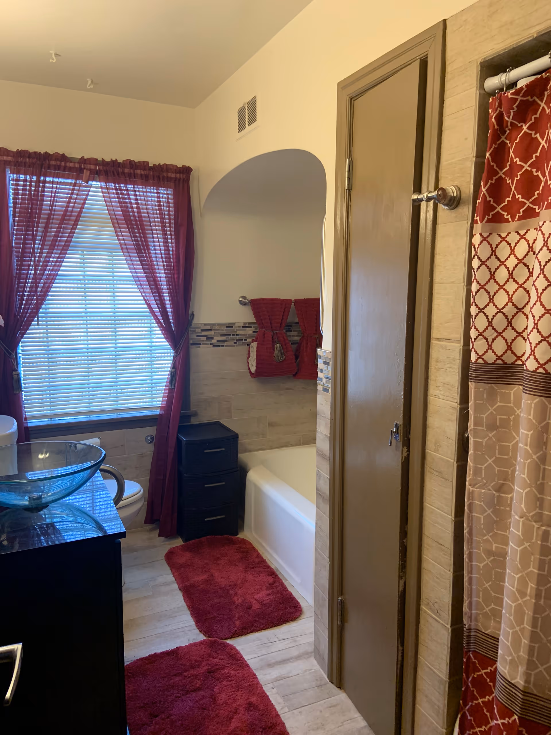 A bathroom with a window covered by red curtains and white blinds. There is a glass vessel sink on a dark vanity, a white toilet, a bathtub with a red and beige patterned shower curtain, and red bath mats on the floor. Red towels are hanging on the wall above the bathtub. The walls are tiled with beige tiles and a decorative mosaic tile border.