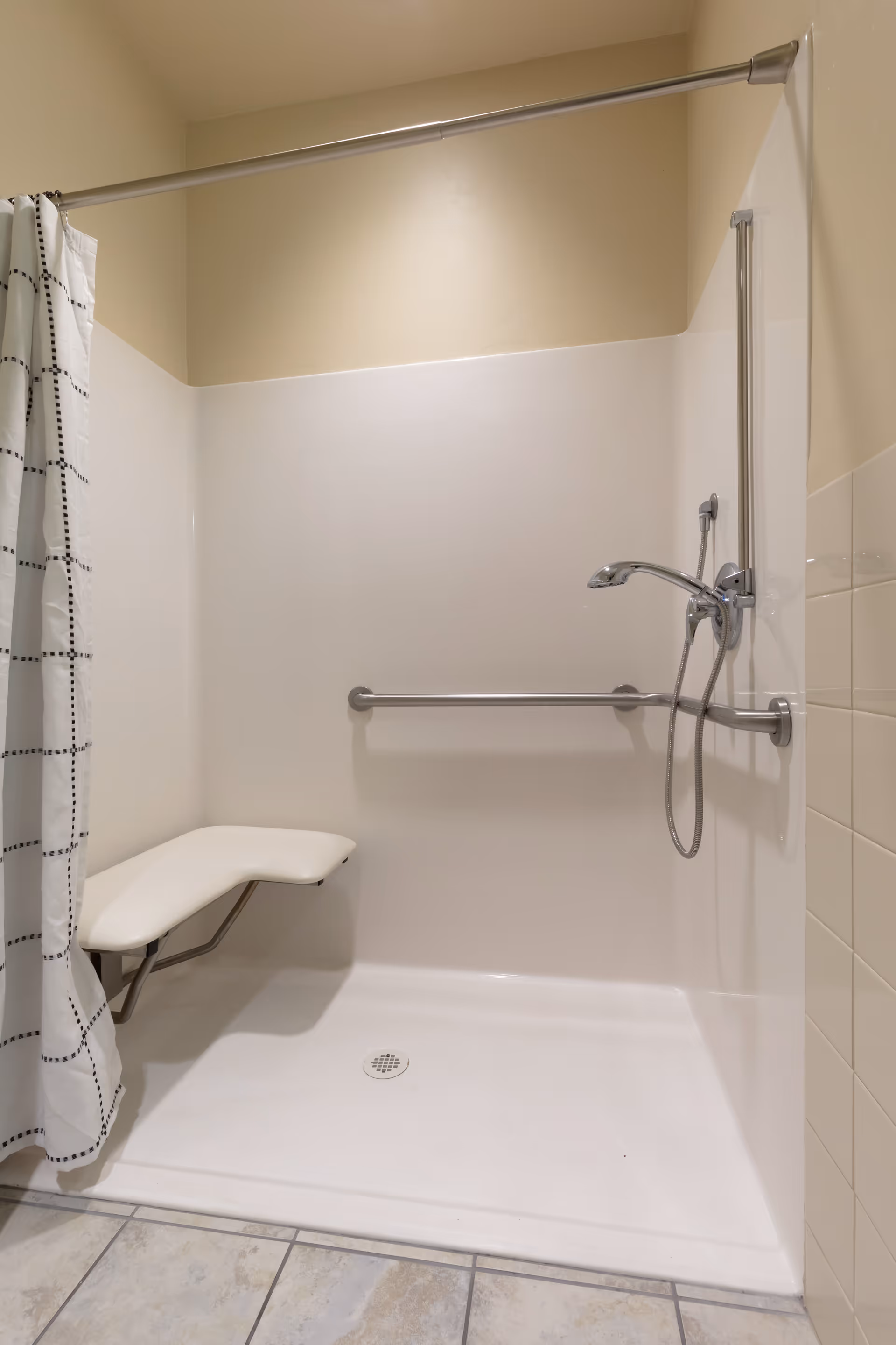 Accessible shower area with a fold-down white seat, a handheld showerhead, a horizontal grab bar, and a white shower curtain with a black grid pattern. The shower floor is white and the walls are a combination of beige paint and white tiles.