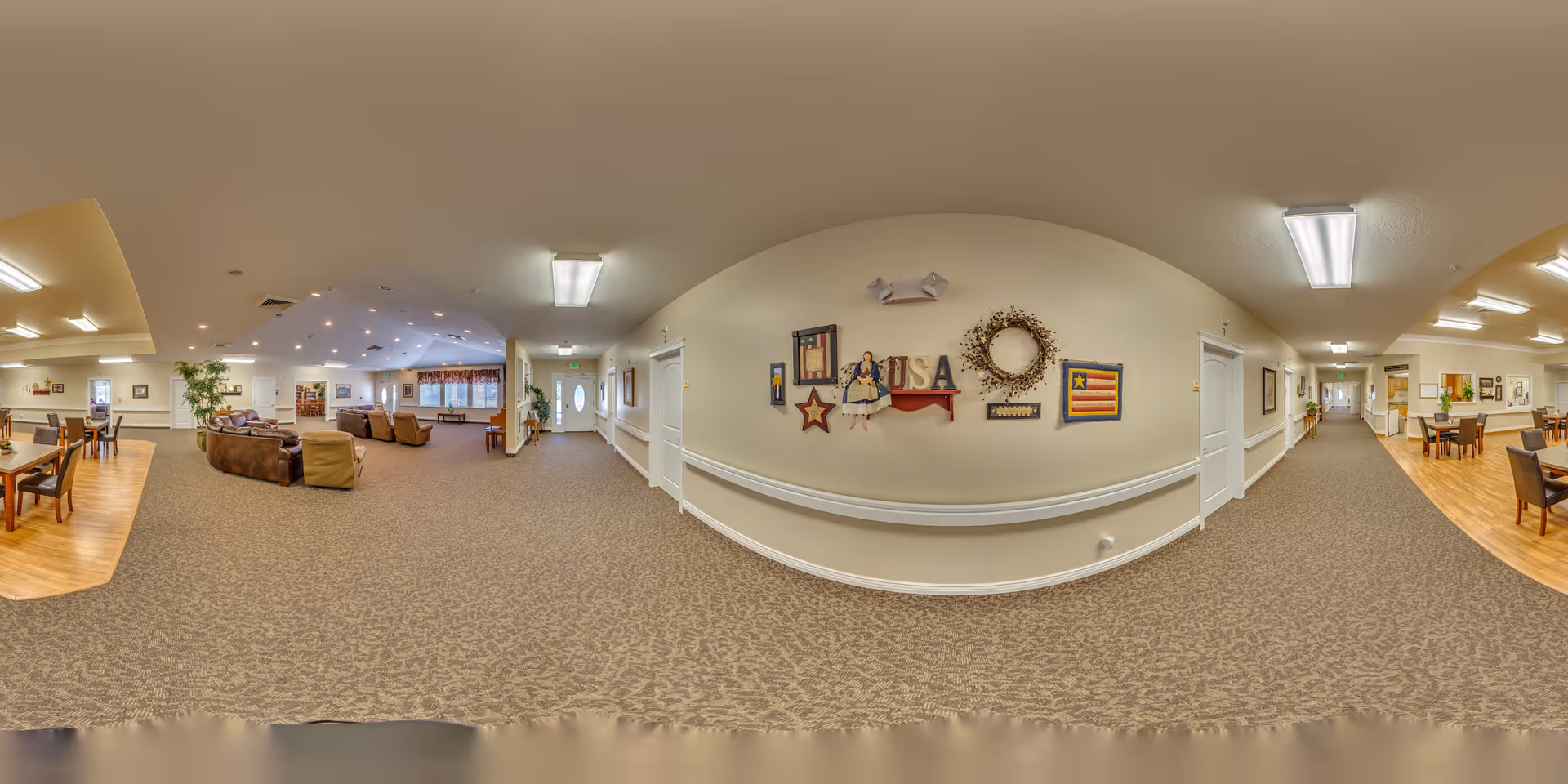 Wide panoramic view of a senior living facility interior showing a spacious common area with carpeted floors and beige walls. The area includes a seating section with brown sofas and armchairs, a dining area with tables and chairs on a wooden floor, and a hallway with doors and wall decorations including patriotic-themed artwork and a wreath. The ceiling has recessed lighting and fluorescent fixtures.