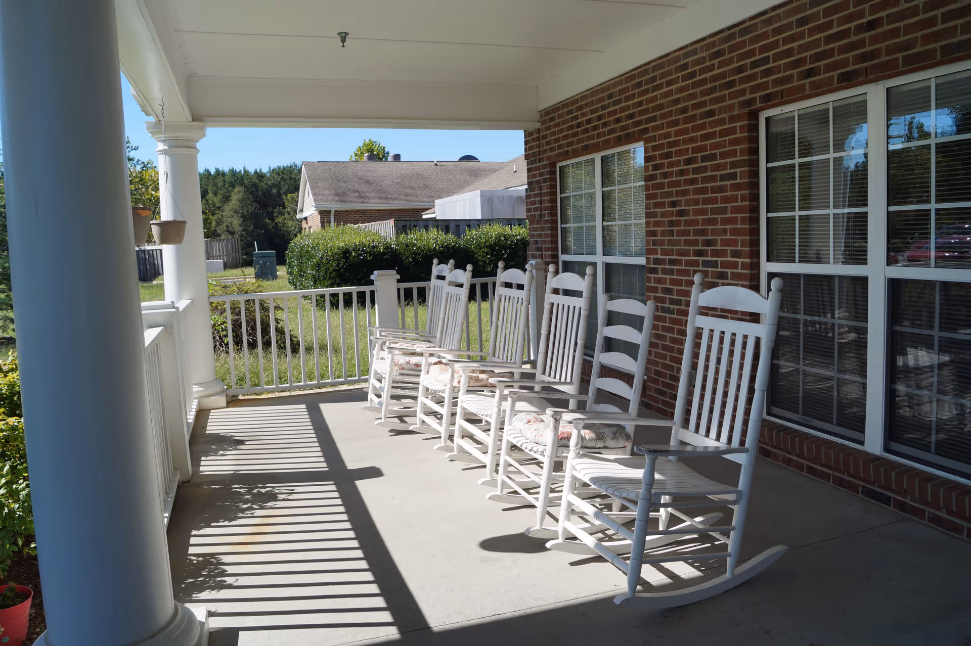 A covered porch with six white wooden rocking chairs lined up against a brick wall with large windows. The porch has white columns and a white railing, with greenery and a neighboring house visible in the background under a clear blue sky.
