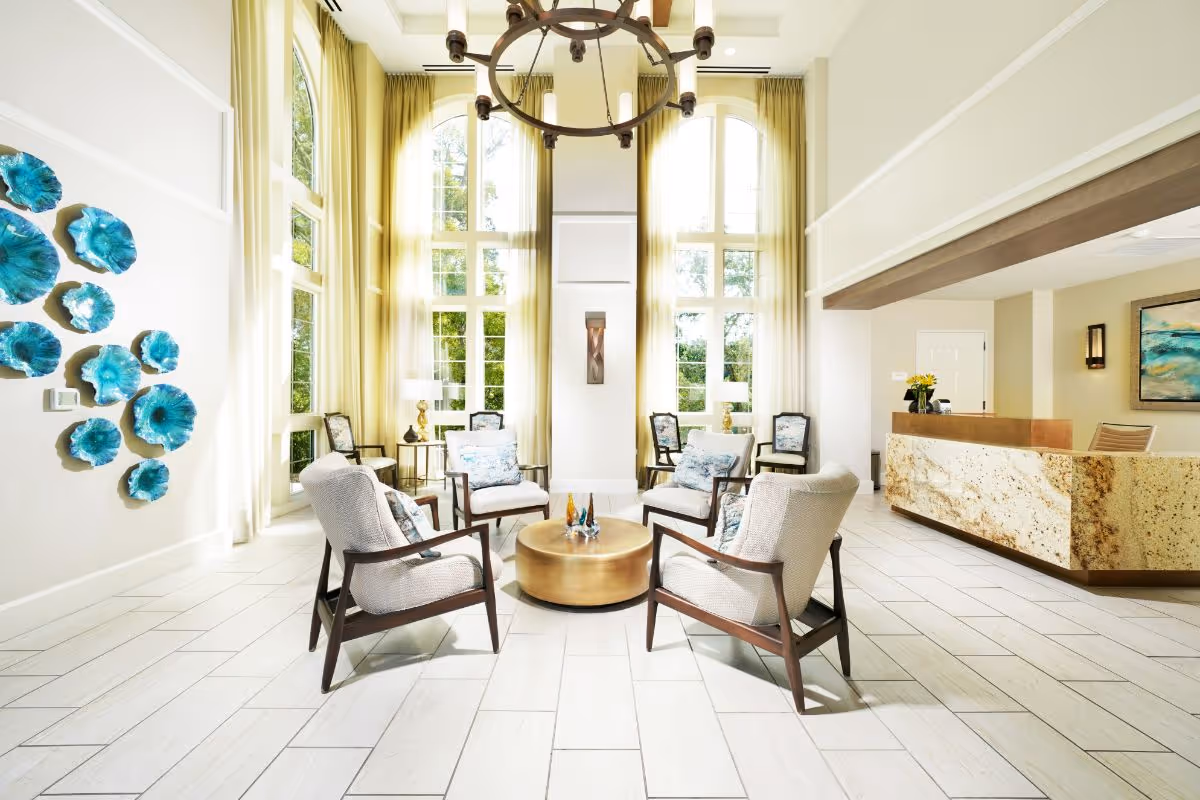 Sunlit lobby with tall arched windows, a central seating arrangement of four armchairs around a round coffee table, and a reception desk to the right.