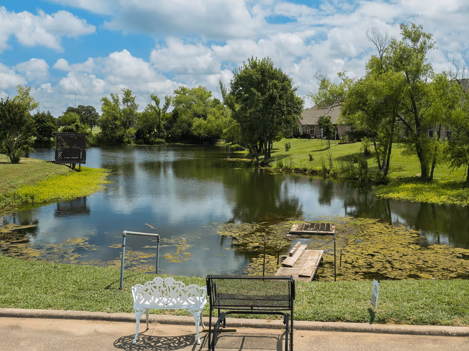 A peaceful outdoor scene at Pegasus Landing of Chisholm Trail featuring a small pond surrounded by green grass and trees under a partly cloudy blue sky. Two benches, one white and one black, are placed on a paved area facing the pond. A small wooden dock extends into the water.