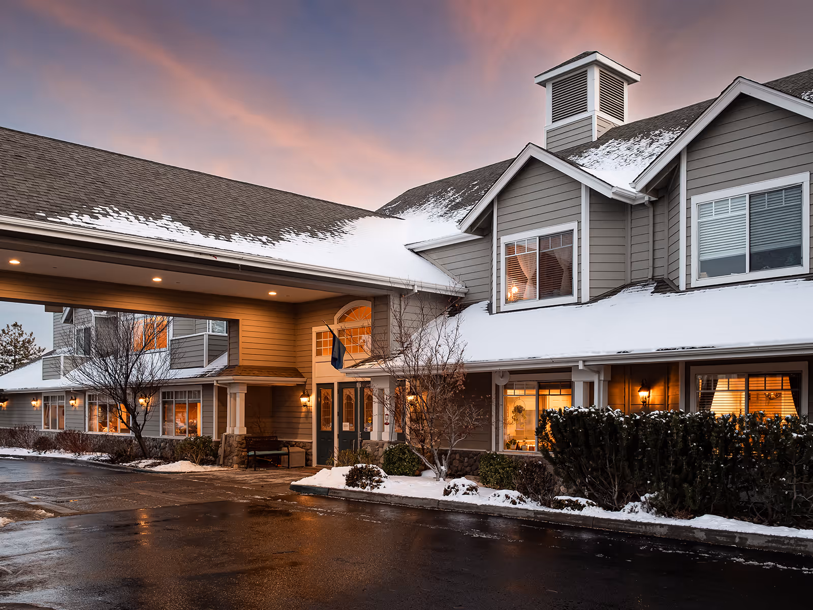 Snow-covered senior living facility exterior with illuminated entrance at sunset.