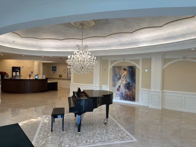 A spacious, elegant lobby area featuring a black grand piano with a matching bench on a decorative rug. Above the piano hangs a large, ornate crystal chandelier. In the background, there is a reception desk with a person seated behind it, and a large painting of a woman in a flowing dress holding a violin is displayed on the wall. The floor is polished tile, and the walls are decorated with white paneling and molding.