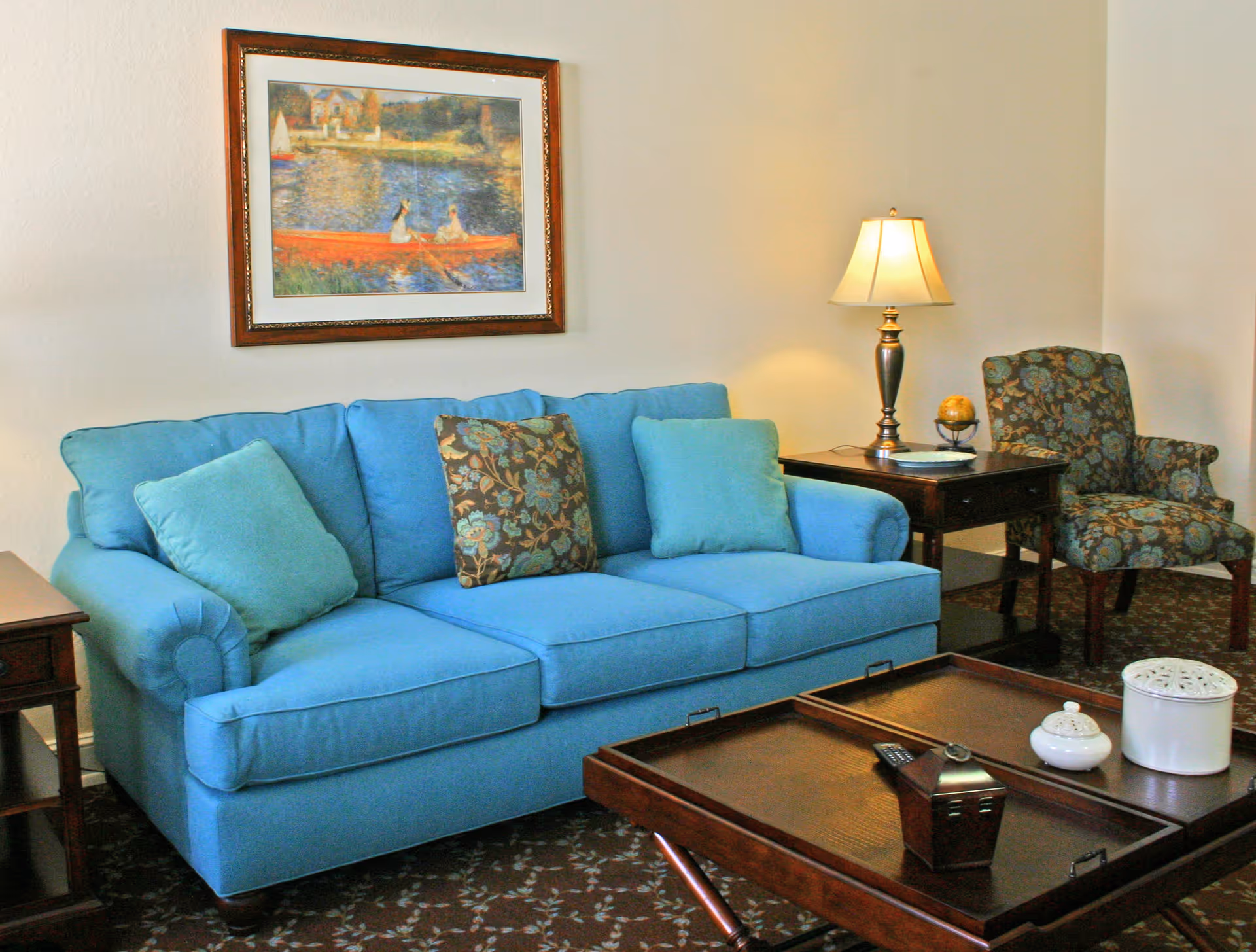 A cozy living room with a blue sofa adorned with three cushions, a wooden coffee table with decorative items, a side table with a lamp and a small globe, and a floral-patterned armchair. A framed painting of a lakeside scene hangs on the wall above the sofa.