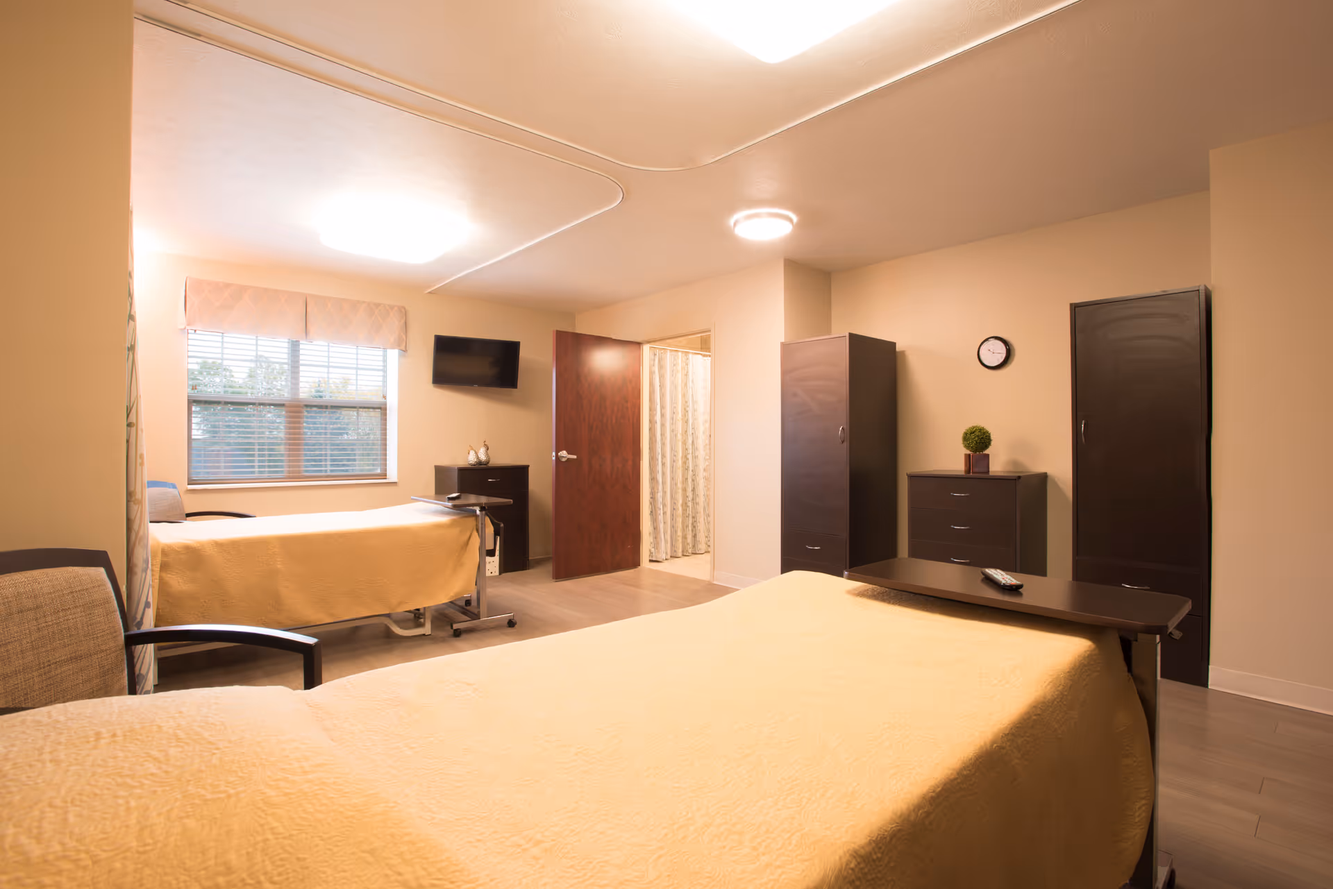 A well-lit room with two beds covered in yellow bedspreads, a chair, a wall-mounted TV, two tall dark wardrobes, a small dresser with a plant, a clock on the wall, and an open door leading to a bathroom with a shower curtain.
