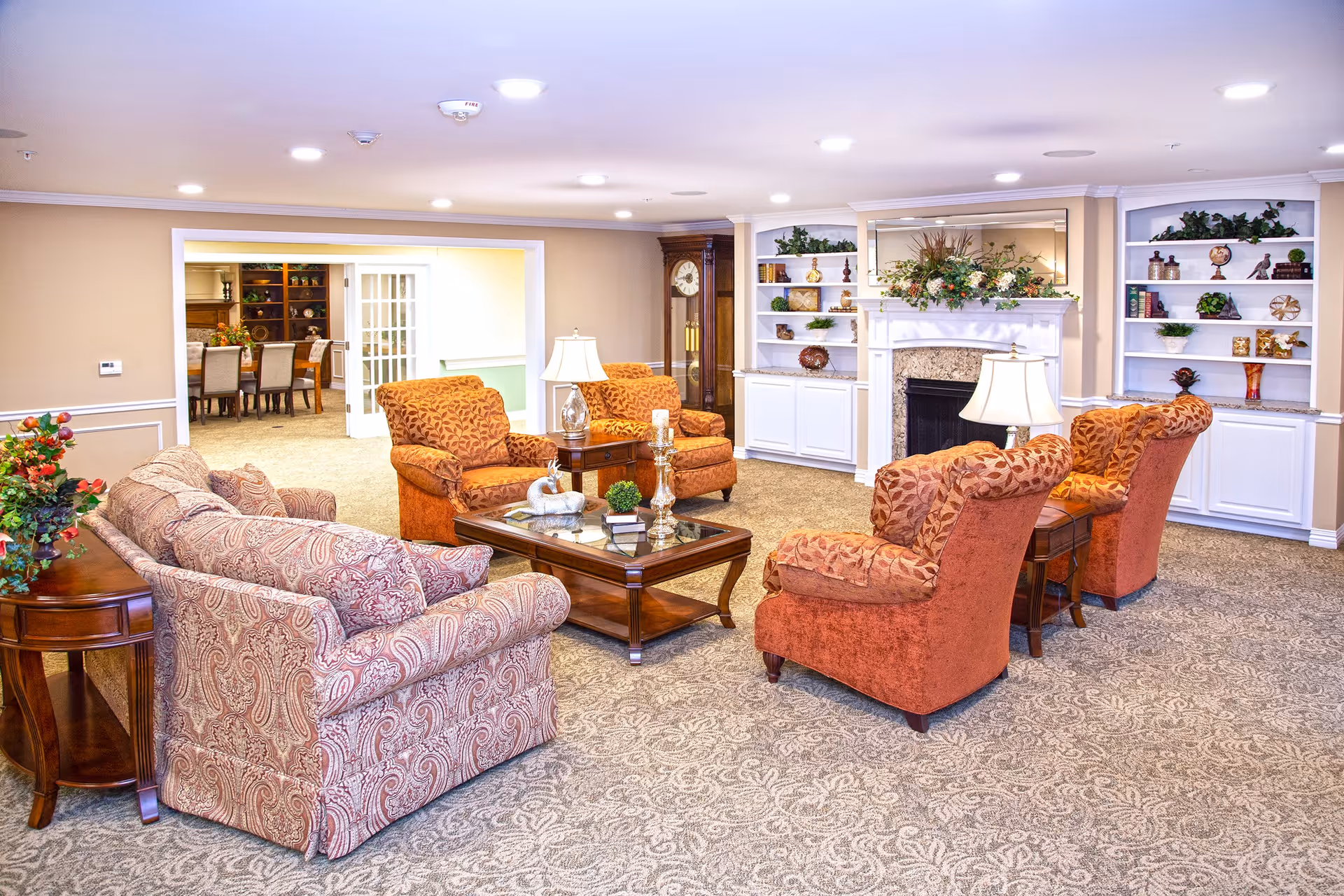 A cozy living room area with patterned carpet, a floral-patterned sofa, four orange armchairs with leaf designs, a wooden coffee table with decorative items, two table lamps, a fireplace with a floral arrangement on the mantel, built-in white shelves with various decorations, and a tall grandfather clock. In the background, there is a dining area with a table and chairs visible through an open doorway.