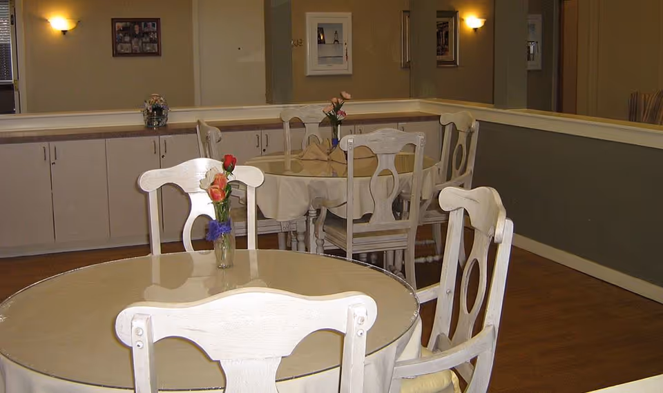 Interior view of a dining area with round tables covered with tablecloths and white wooden chairs. Each table has a small vase with flowers. The room has beige walls, wooden flooring, and soft wall lighting. Cabinets line the back wall with framed pictures hanging above.