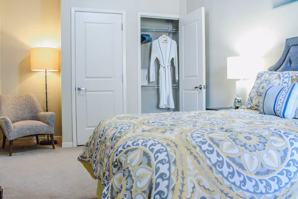 Bright bedroom featuring a patterned bed with decorative pillows, an open closet displaying a hanging white robe, an armchair, and lamps.