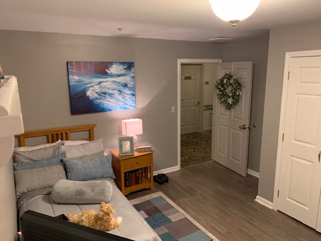 A cozy bedroom in a senior living facility with a bed covered in gray and blue bedding, a small wooden nightstand with a lamp and framed photo, a painting of ocean waves on the wall, an open door with a wreath hanging on it, and a small rug on the wooden floor. A cat is lying on the bed near the footboard.