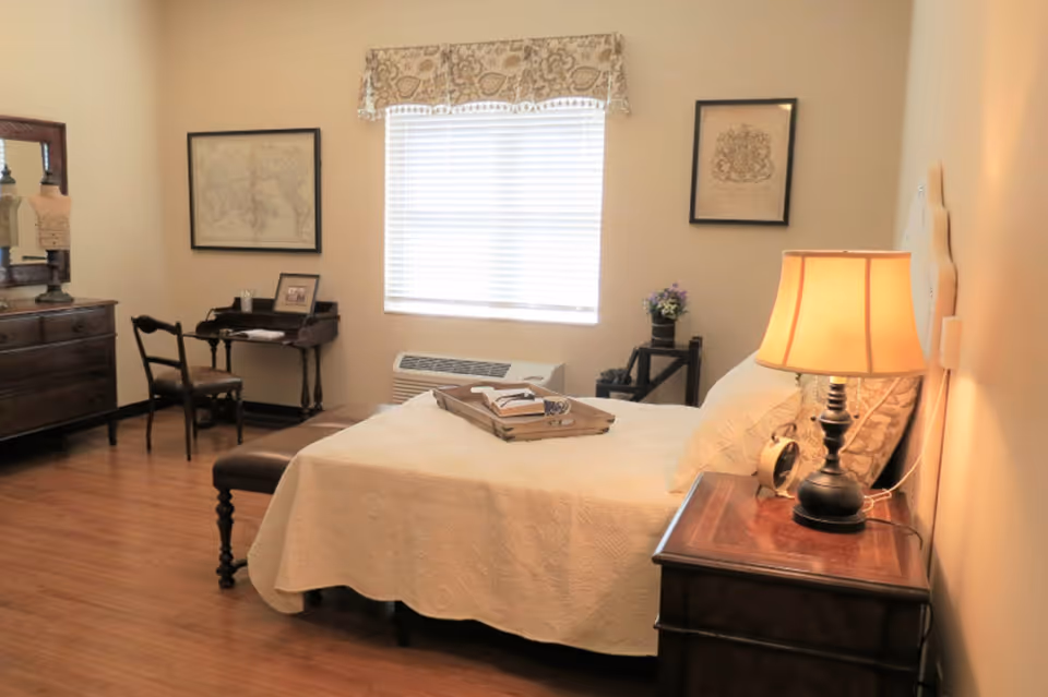 A cozy bedroom with a neatly made bed covered in a white quilt. There is a wooden nightstand with a lamp and an alarm clock next to the bed. A wooden bench is placed at the foot of the bed. The room has wooden flooring, a window with blinds and a patterned valance, and two framed pictures on the walls. A small desk with a chair and a dresser with a mirror are also visible.