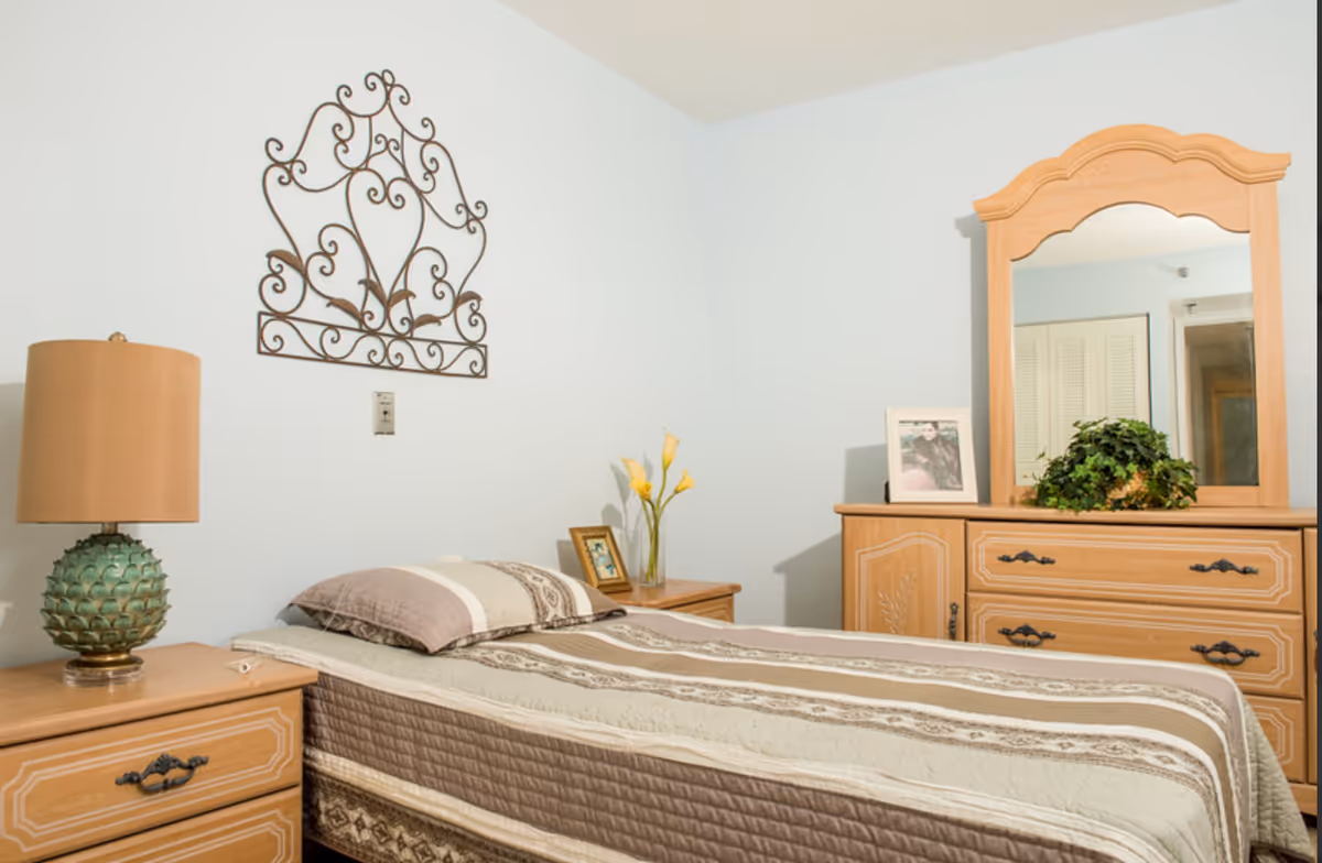 A bedroom with a single bed covered in a striped quilt in shades of beige and brown. Next to the bed is a wooden nightstand with a green pineapple-shaped lamp. On the wall above the bed is a decorative wrought iron wall art. Across from the bed is a wooden dresser with a large mirror, a framed photo, and a potted plant on top. The walls are painted light blue.