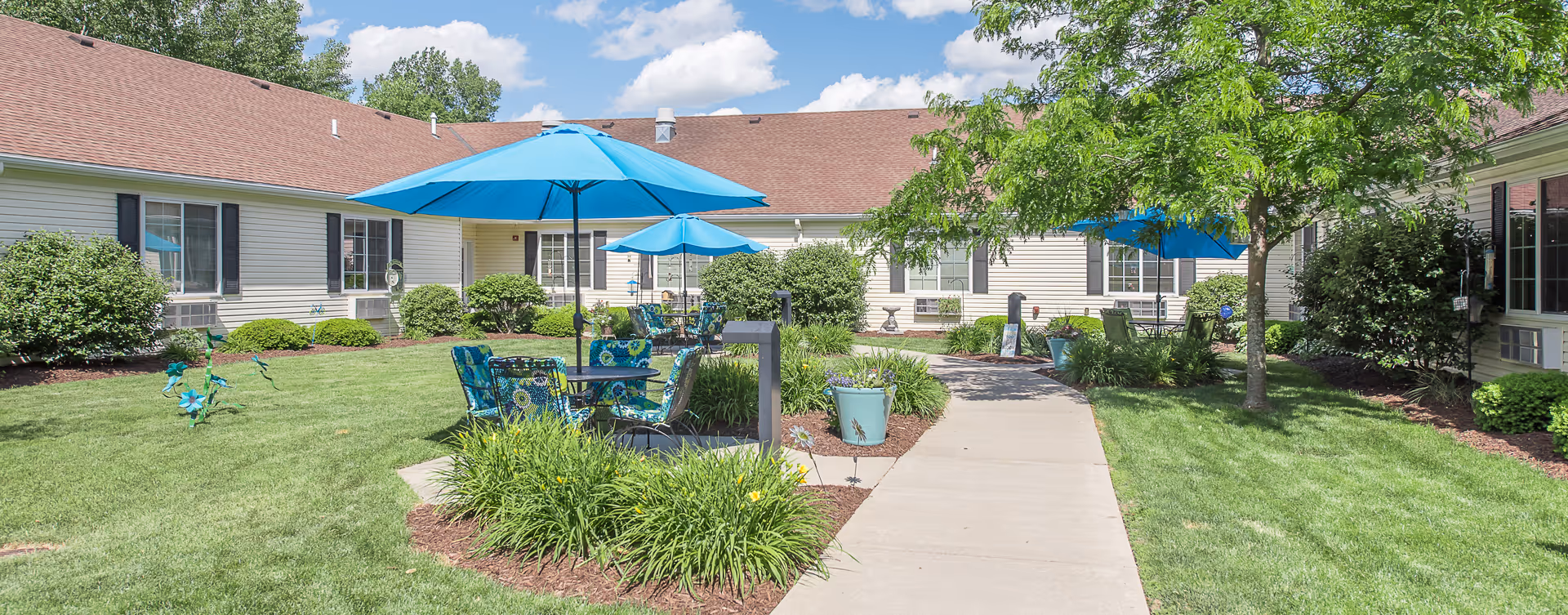 Outdoor courtyard area at Bickford of Midland with green grass, shrubs, and trees. There are several blue patio umbrellas shading tables and chairs arranged on the lawn. A concrete walkway runs through the courtyard, surrounded by flower beds and potted plants. The building with beige siding and brown roof encloses the courtyard.