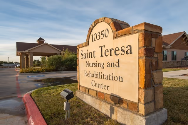 Stone sign at the entrance of Saint Teresa Nursing and Rehabilitation Center with the address number 10350, situated on a grassy area near a driveway and the facility building in the background under a clear sky.