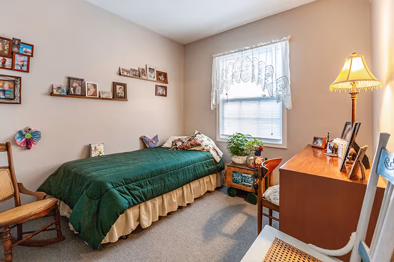 Small furnished bedroom with a single bed, desk, window, and personal photos on the wall.
