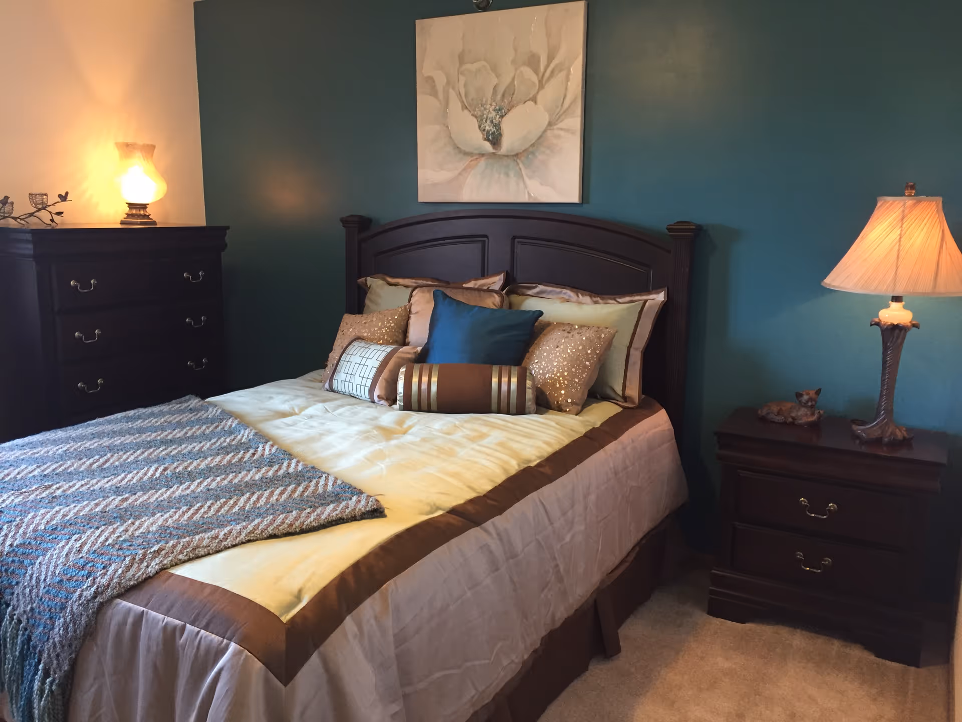 A cozy bedroom with a dark wooden bed frame and a neatly made bed featuring multiple decorative pillows and a folded blanket at the foot. The walls are painted teal, and there is a large floral painting above the bed. On the left side, there is a dark wooden dresser with a glowing lamp on top, and on the right side, a matching nightstand with a lit lamp and a small decorative figurine.