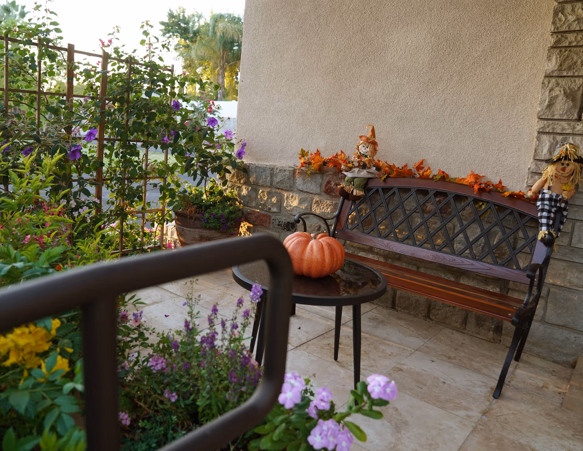 A cozy outdoor patio area with a metal and wood bench decorated with autumn leaves and two scarecrow dolls. A small round table in front of the bench holds a large orange pumpkin. Surrounding the patio are various green plants and flowers, with a trellis supporting climbing plants in the background.